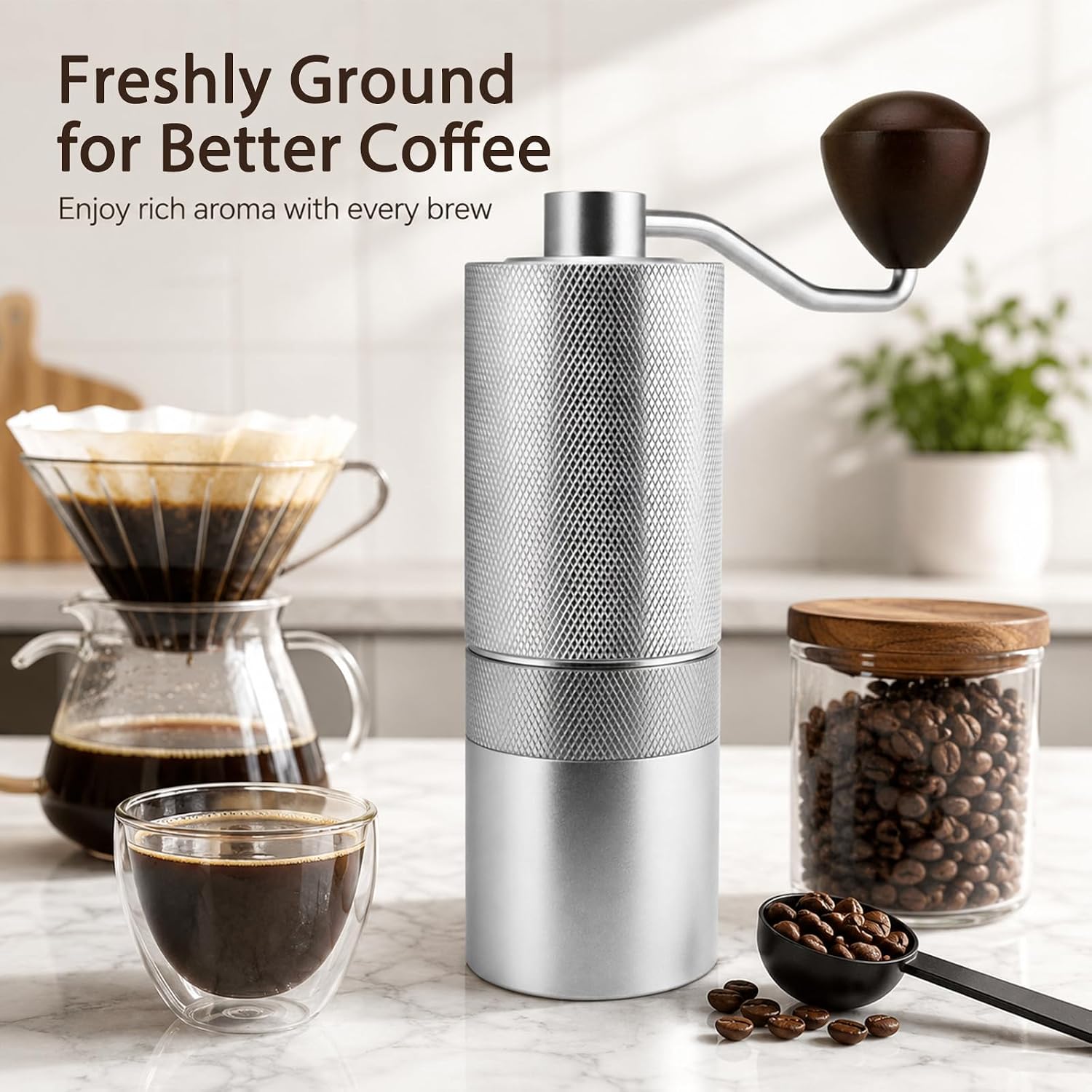 A manual coffee grinder with a textured silver body and a dark brown wooden handle is shown on a marble countertop, surrounded by coffee beans in a glass jar, brewed coffee in a pour-over dripper and glass cup, and a black scoop. Text on the top left reads 'Freshly Ground for Better Coffee'.
