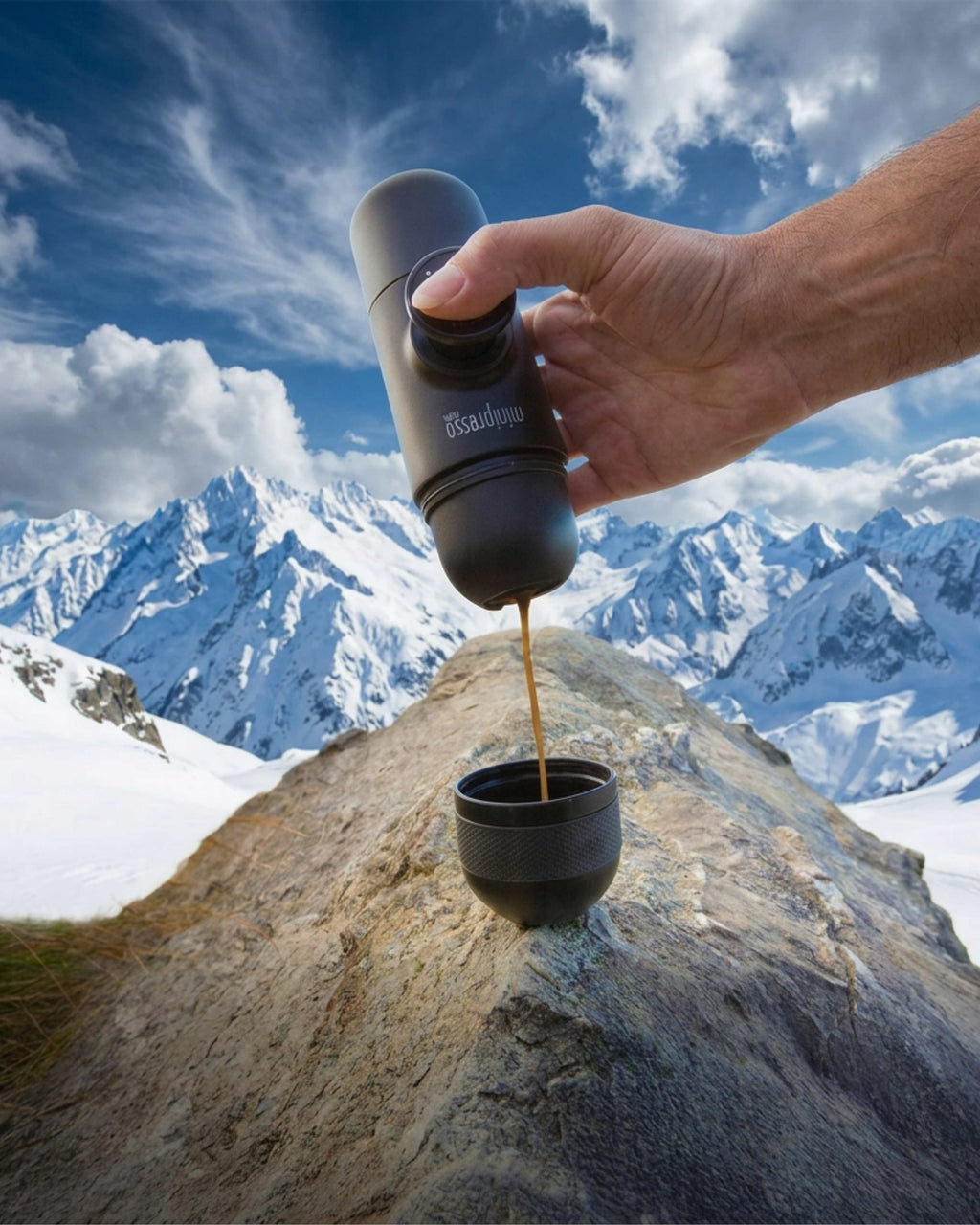 A hand holds a black portable coffee maker above a small cup, pouring coffee, with a snowy mountain landscape and blue sky in the background. The product brand 'Brewery' is visible on the device.