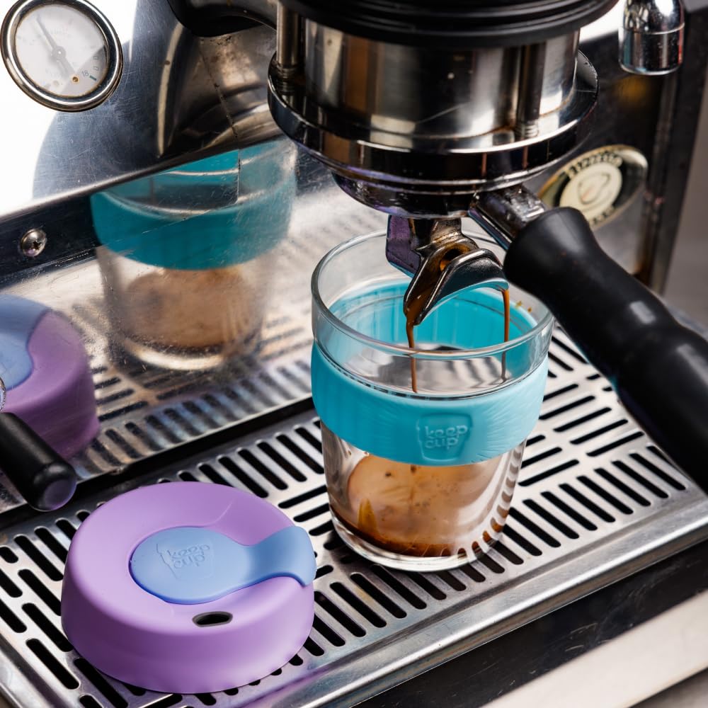 A clear glass cup with a bright turquoise silicone sleeve sits under a stainless steel espresso portafilter, with espresso dripping into the cup. A matching purple and turquoise lid lies on the metal drip tray nearby.