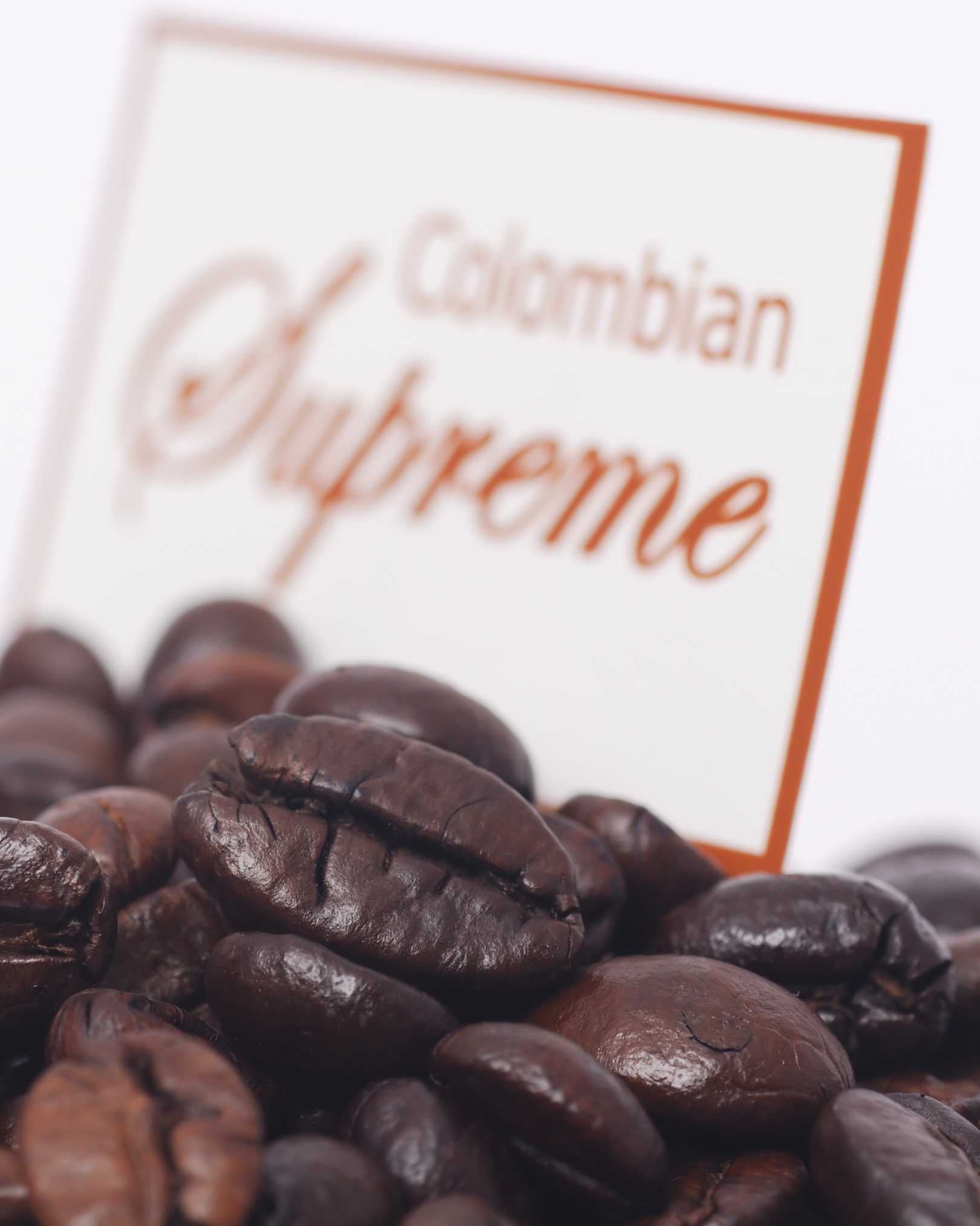 A close-up of roasted coffee beans in the foreground with a blurred white sign reading 'Colombian Supreme' in the background.