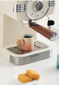 A close-up of a cream-colored Schmieder espresso machine with a wooden-handled steam wand, dispensing coffee into a pink-to-blue gradient ceramic cup. The Schmieder brand name is visible on the silver base trim, and two golden-brown cookies sit on a small white plate in the foreground.