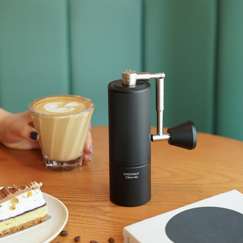 Black coffee grinder on a wooden table with a glass of coffee and a slice of cake.