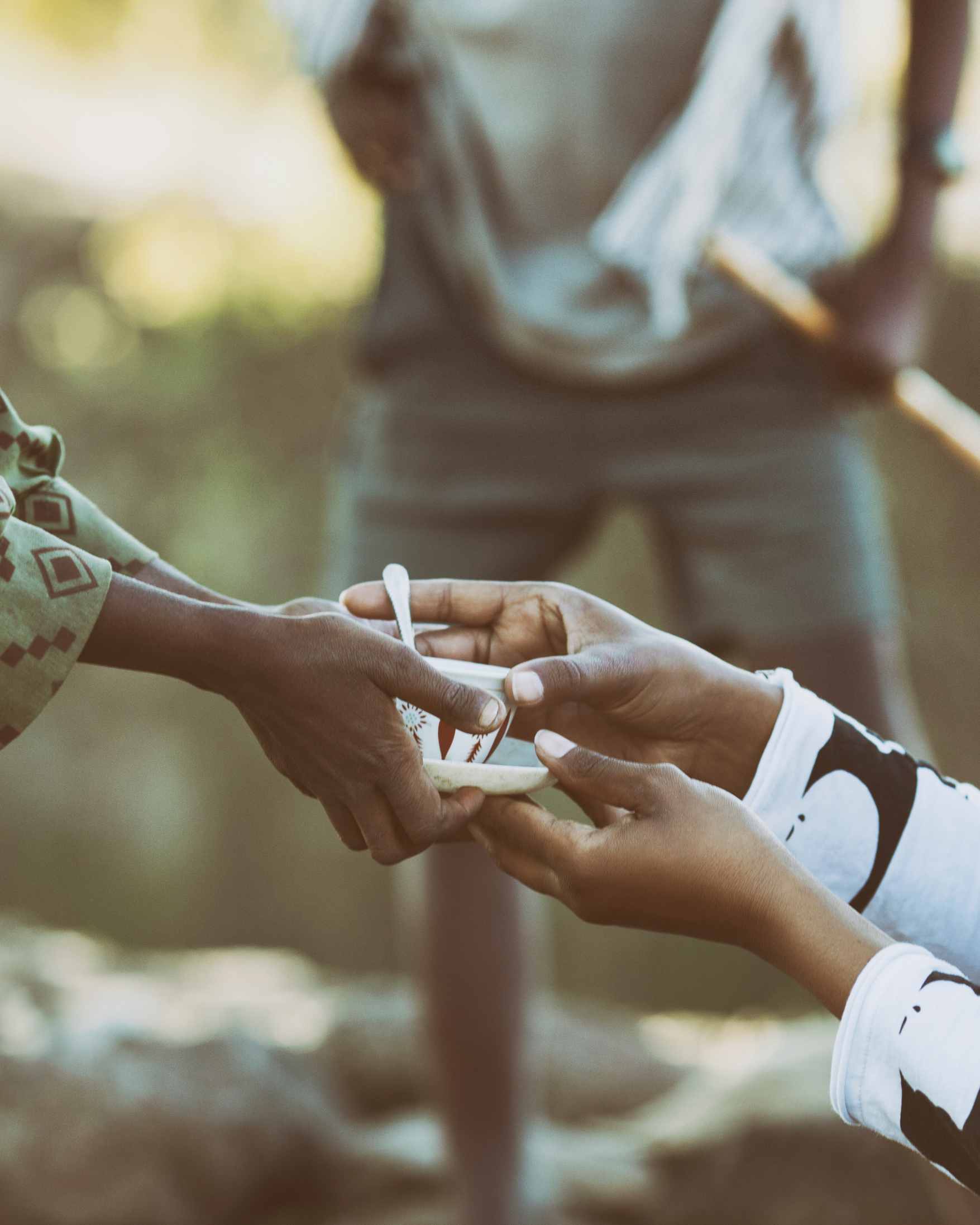 A close-up lifestyle shot of hands exchanging a small white ring box or container outdoors, with a blurred person standing in the background.