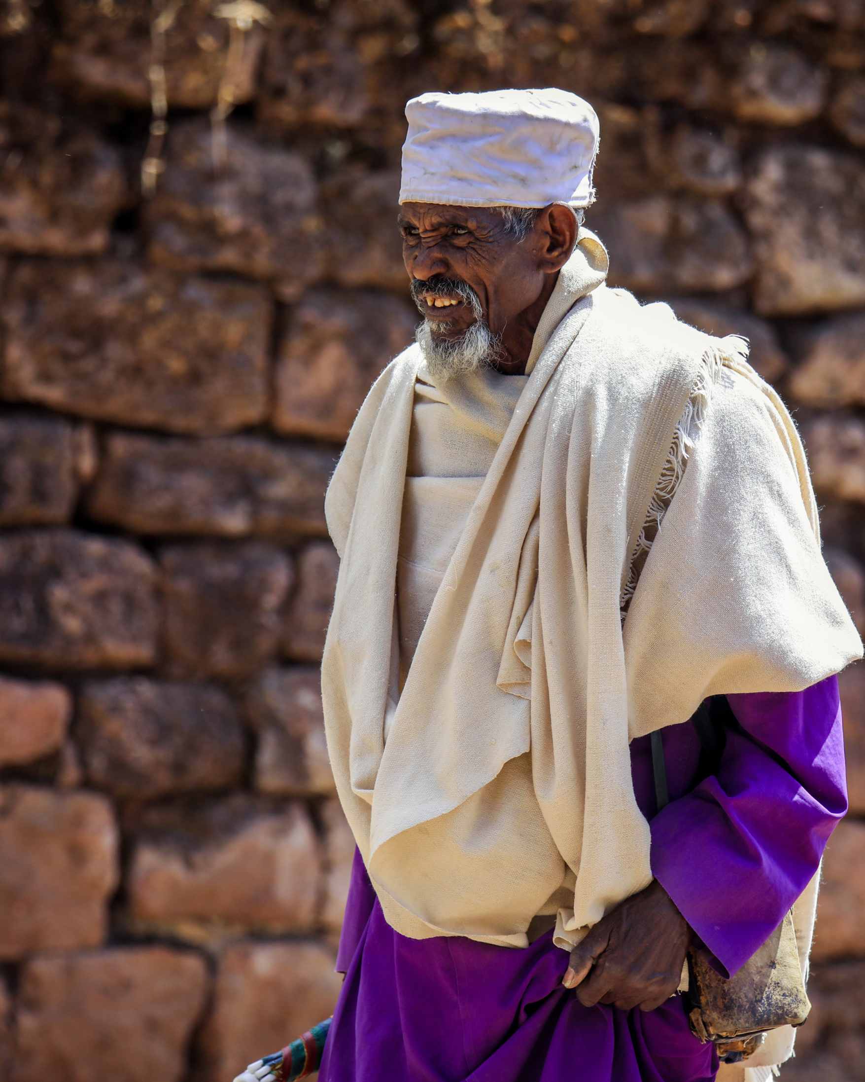 An elderly man wearing traditional Ethiopian clothing, including a white hat, a cream-colored shawl over a white tunic, and a purple skirt, standing in front of a rustic stone wall.