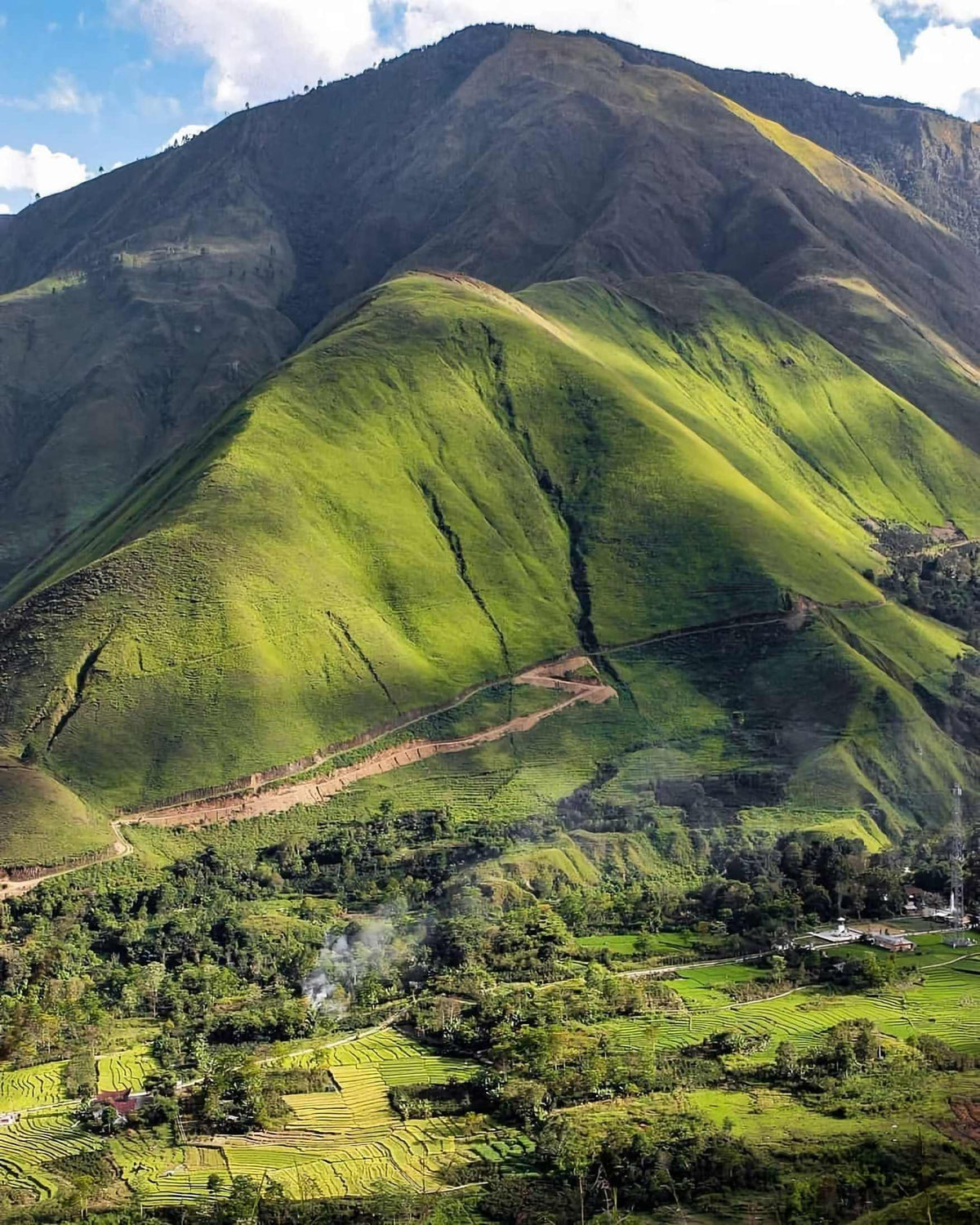 A scenic landscape photograph of a lush green mountain with terraced fields and small buildings in the valley below, under a blue sky with clouds.