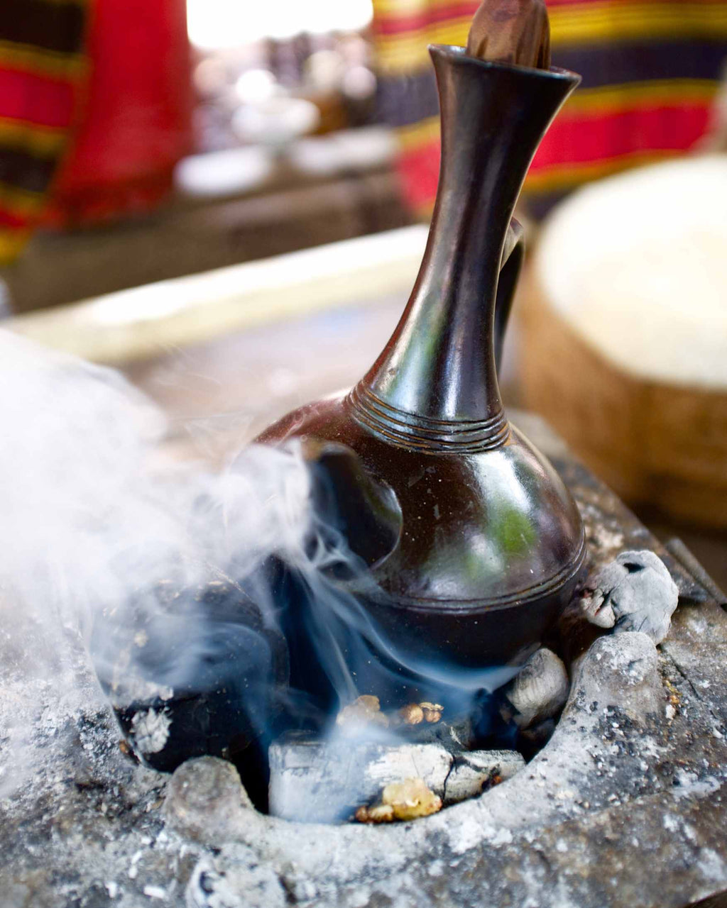 A dark metallic ceremonial or decorative vessel with a flared neck and curved base, shown sitting in a smoky stone burner or fire pit. The background is softly blurred with warm red, yellow, and brown tones.