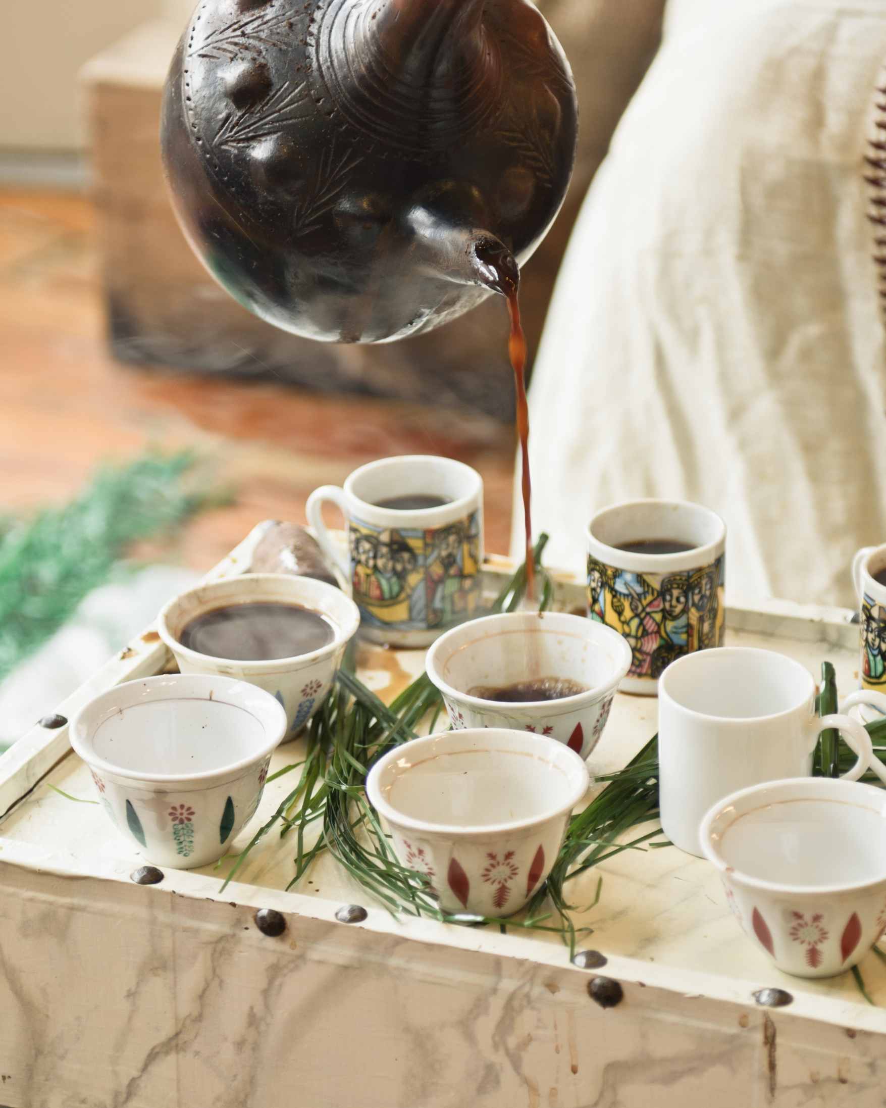 A dark metal teapot pouring dark coffee into a row of small white ceramic cups decorated with colorful floral and geometric patterns, arranged on a marble-patterned tray with green sprigs.