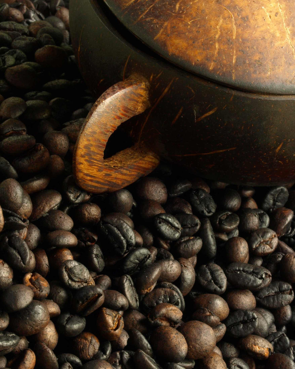 A close-up of roasted coffee beans surrounding a dark brown coconut shell teapot with a wooden handle.