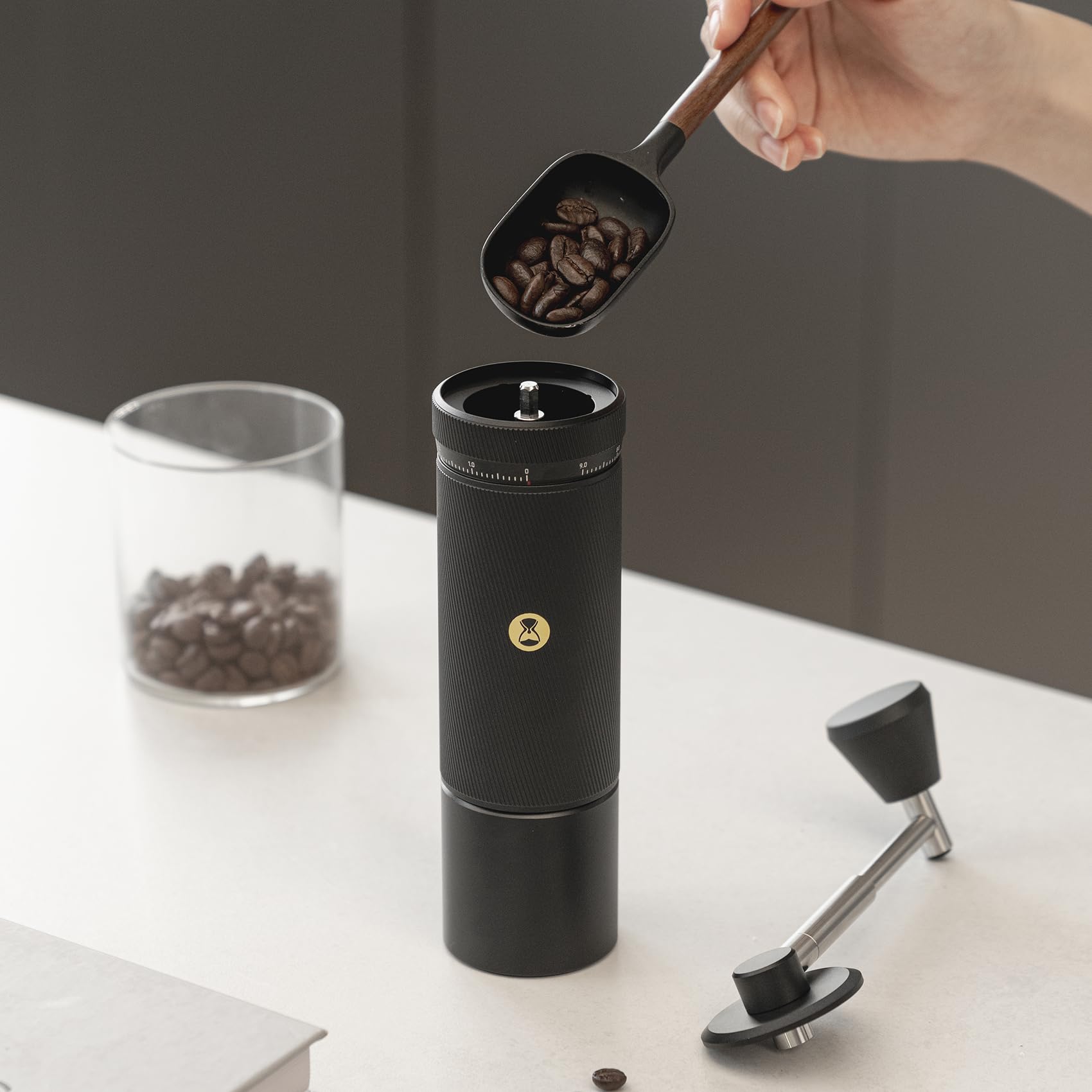 Person adding to a black manual coffee grinder on a light surface.