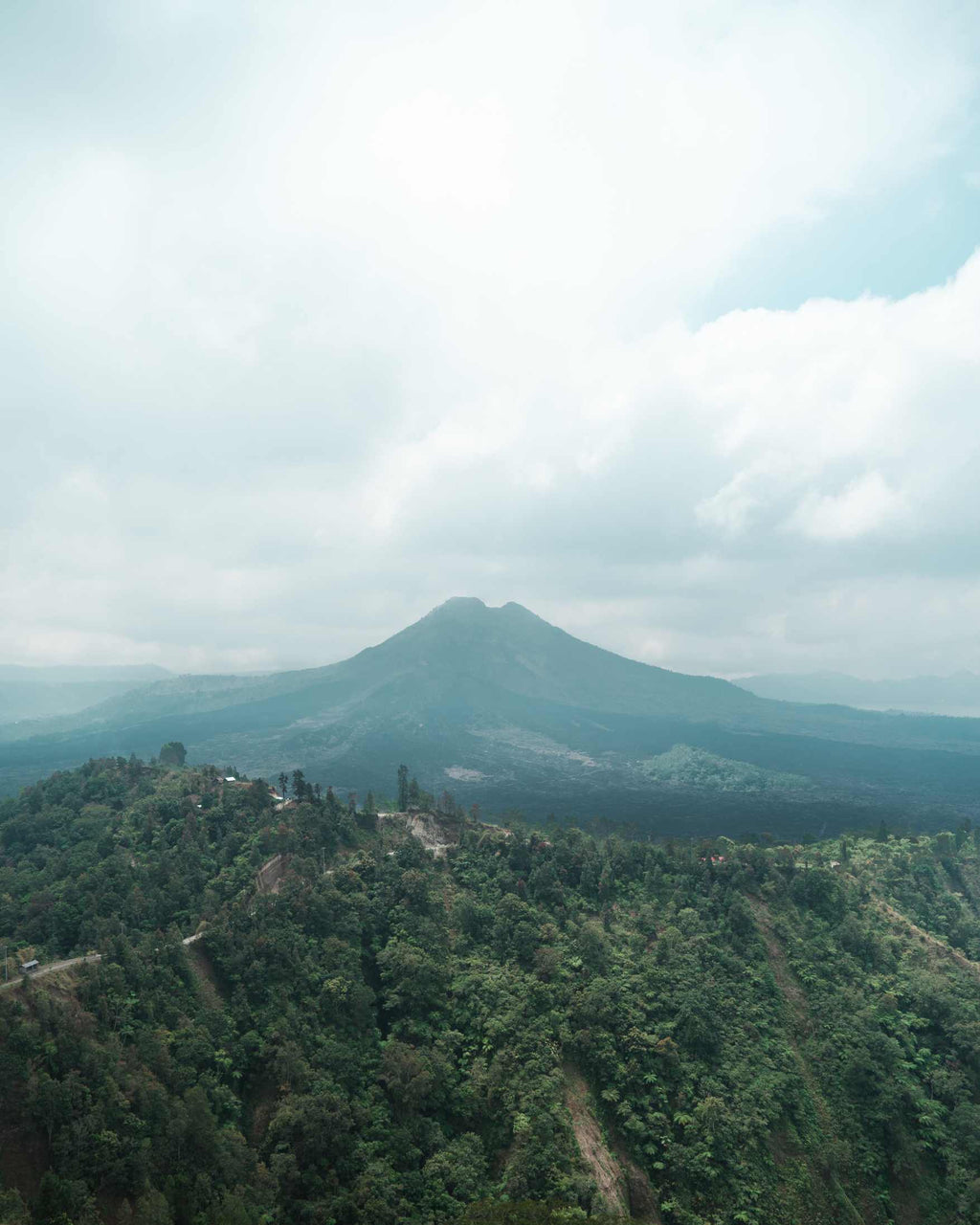 A scenic landscape photograph of a mountain peak rising above a densely forested green hillside under a cloudy sky.