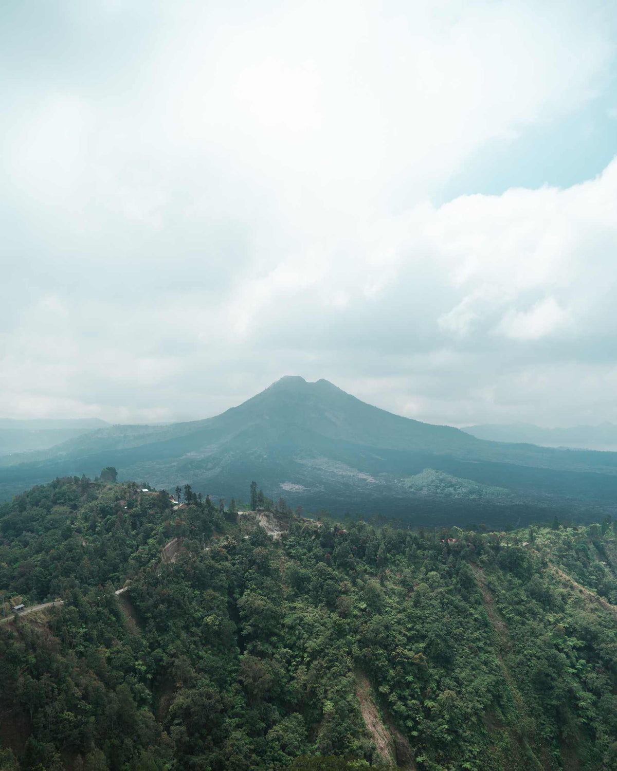 A scenic landscape photograph of a mountain peak rising above a densely forested green hillside under a cloudy sky.