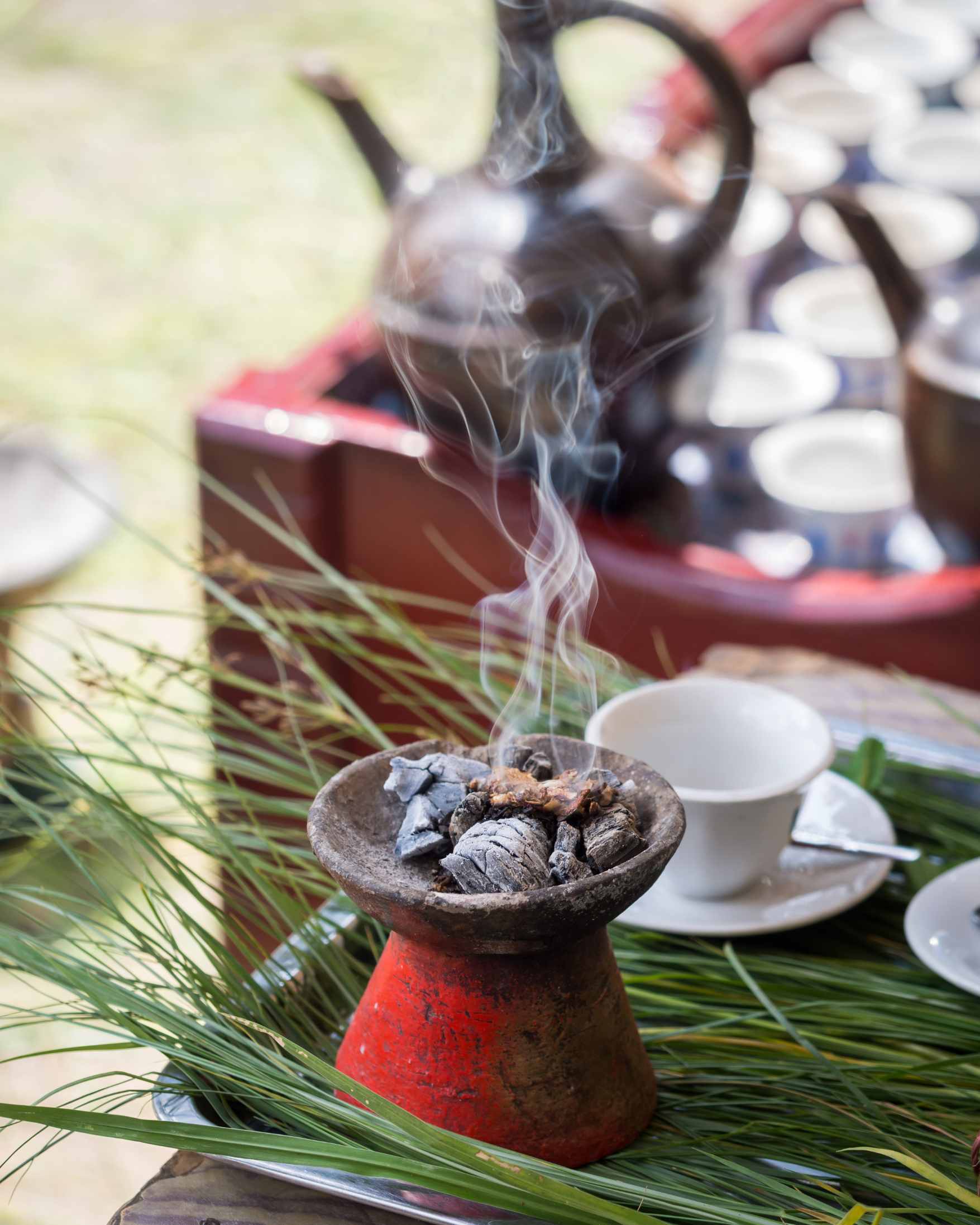 A traditional clay incense burner with glowing embers and rising smoke, set on a tray with green leaves, a white teacup, and a blurred teapot in the background.