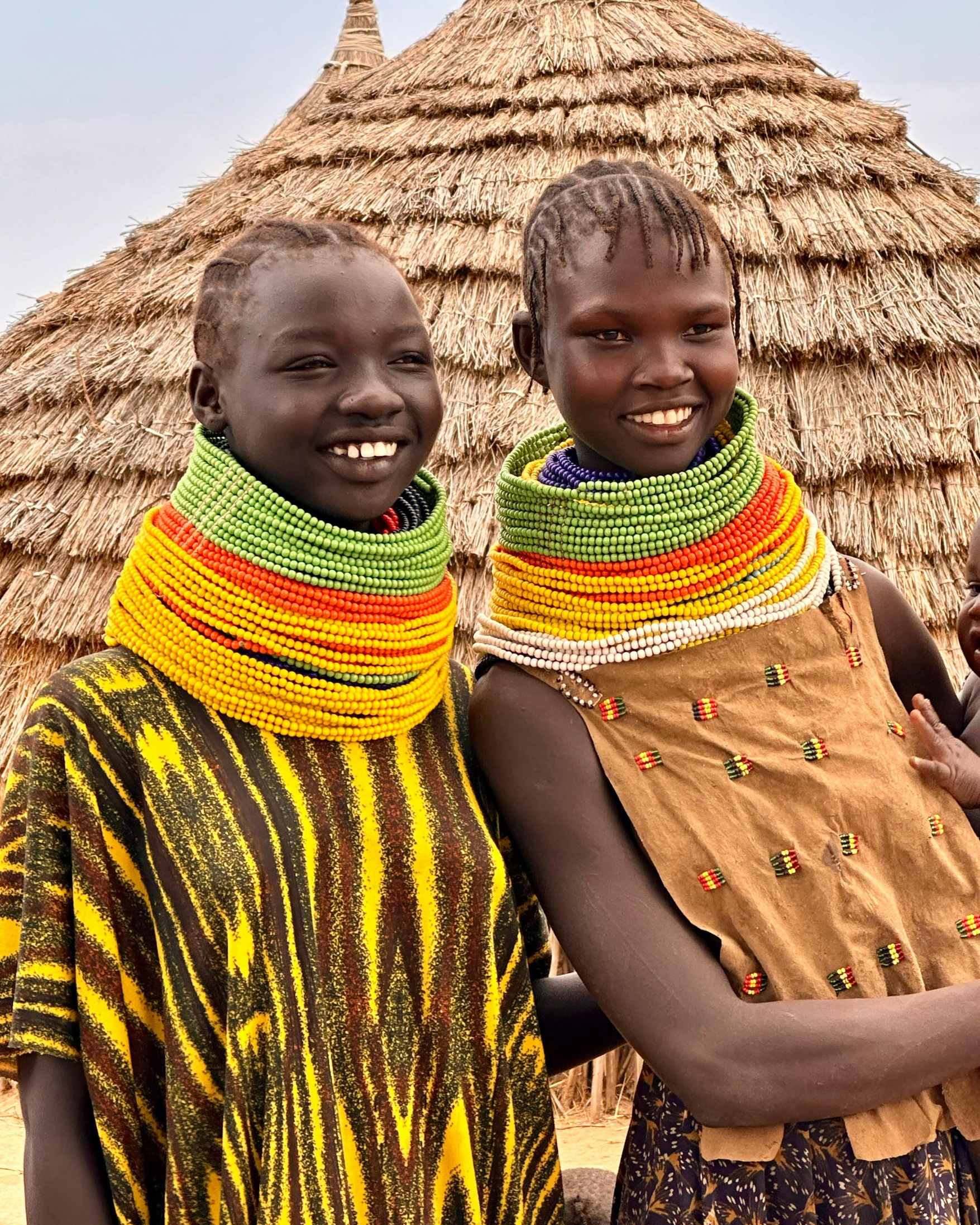 Two smiling young girls standing outdoors in front of a thatched hut, wearing colorful beaded necklaces and patterned clothing. One wears a yellow, brown, and black striped garment, and the other wears a tan top with small decorative beads.