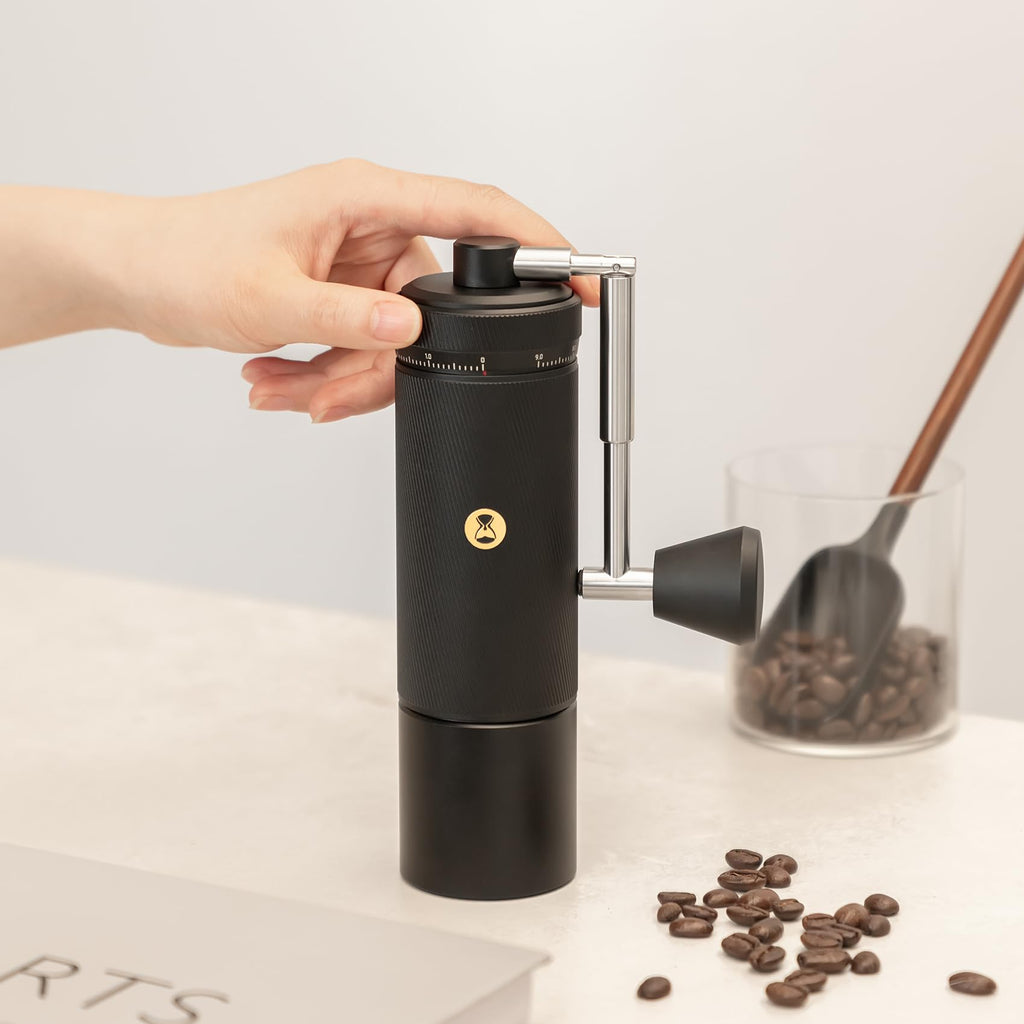 Hand holding a black manual coffee grinder with coffee beans on a light surface.