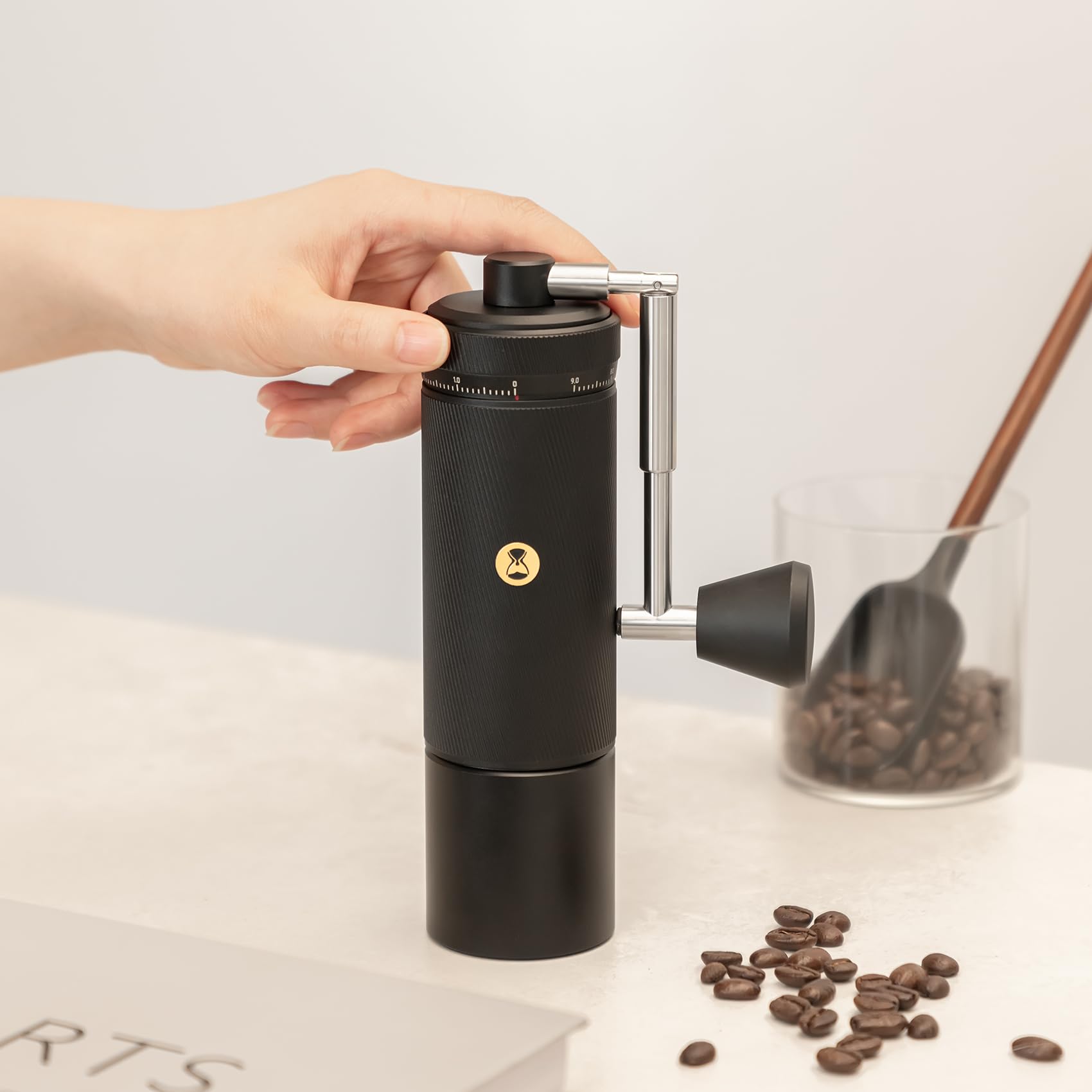 Hand holding a black manual coffee grinder with coffee beans on a light surface.