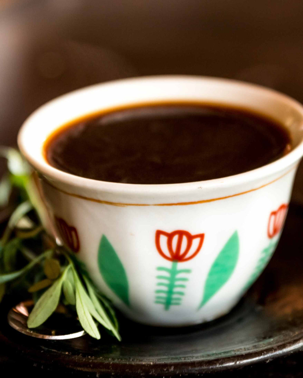 A white ceramic cup filled with dark coffee, decorated with red tulips and green leaves, resting on a dark plate with green herbs in the foreground.