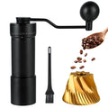 Manual coffee grinder set with a black hand-crank grinder, small cleaning brush, gold-toned conical coffee filter, and a wooden spoon of coffee beans on a white background.