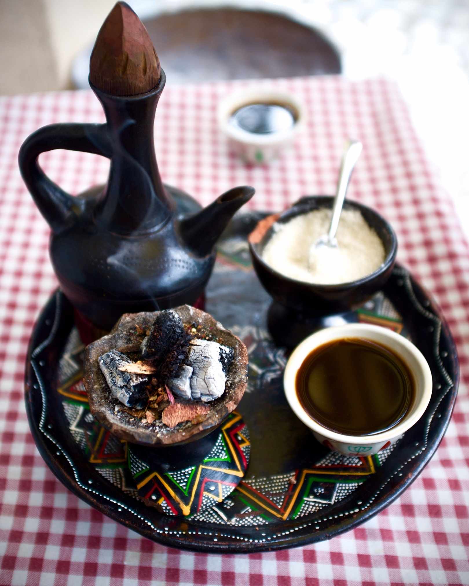 A traditional Moroccan tea set on a red-and-white checkered tablecloth, featuring a black clay teapot, a small bowl of white powder with a spoon, a bowl of dark coffee, and a clay pot with embers on a patterned tray.