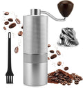 A manual coffee grinder with a textured silver metal body, dark brown wooden handle, and black cleaning brush, shown with scattered coffee beans, ground coffee, and a metal blade on a white background.