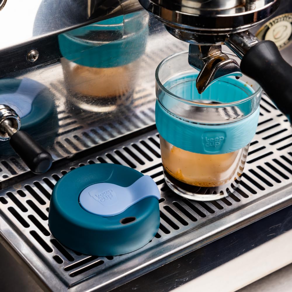 A teal and light blue silicone coffee accessory with a matching lid sits on a stainless steel espresso machine drip tray next to a glass cup with a silicone sleeve under espresso flow.