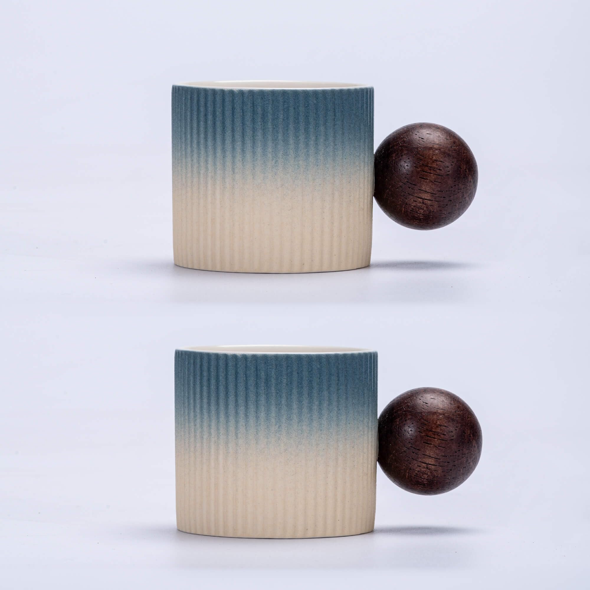 Two ceramic cups with a blue-to-beige ombre gradient and a dark brown wooden sphere handle, shown against a light gray background.