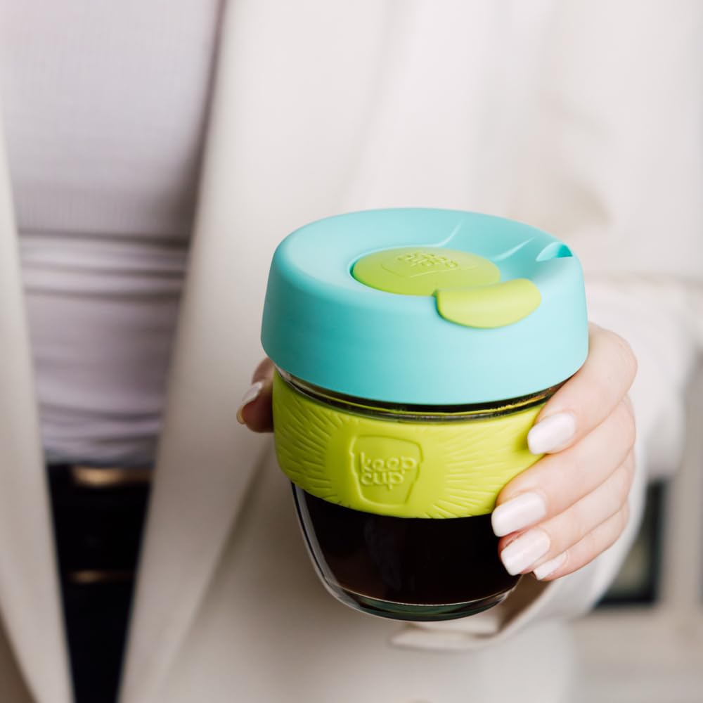 A person holding a reusable glass coffee cup with a bright turquoise lid and a lime green silicone sleeve. The cup is filled with dark coffee, and the person's hand and torso are visible against a soft, light background.